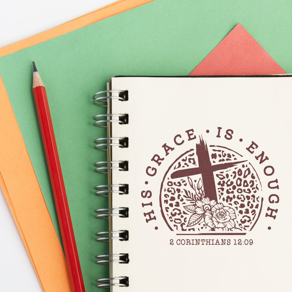 Red pencil beside spiral notebook with brown seal stamp reading His Grace Is Enough on green and orange paper.