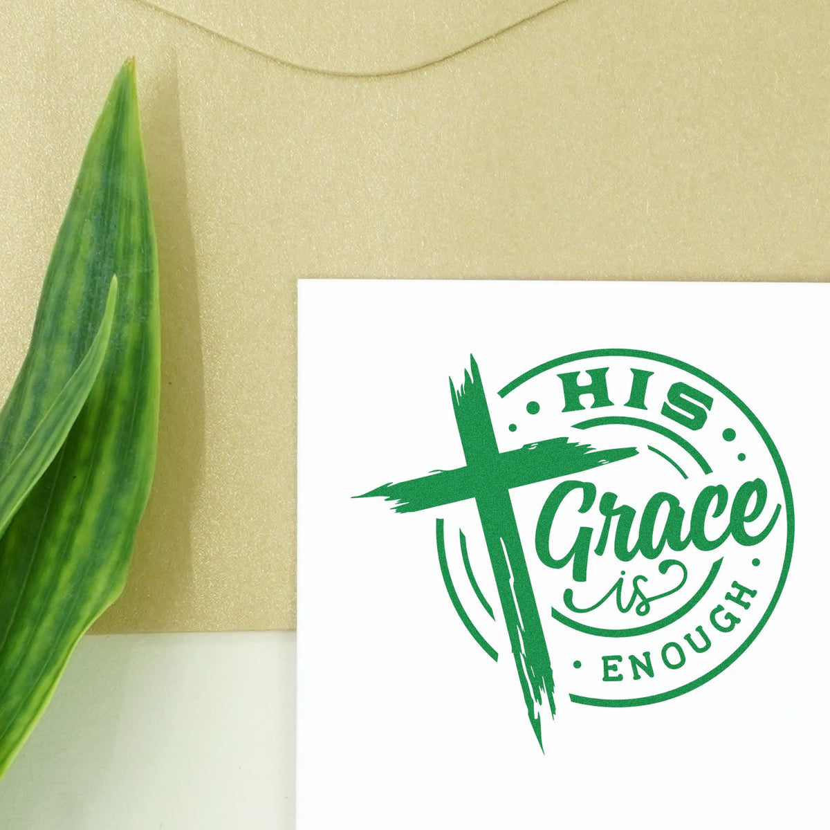 Green stamp and seal design reading His Grace is Enough with a cross on white paper beside a green leaf.