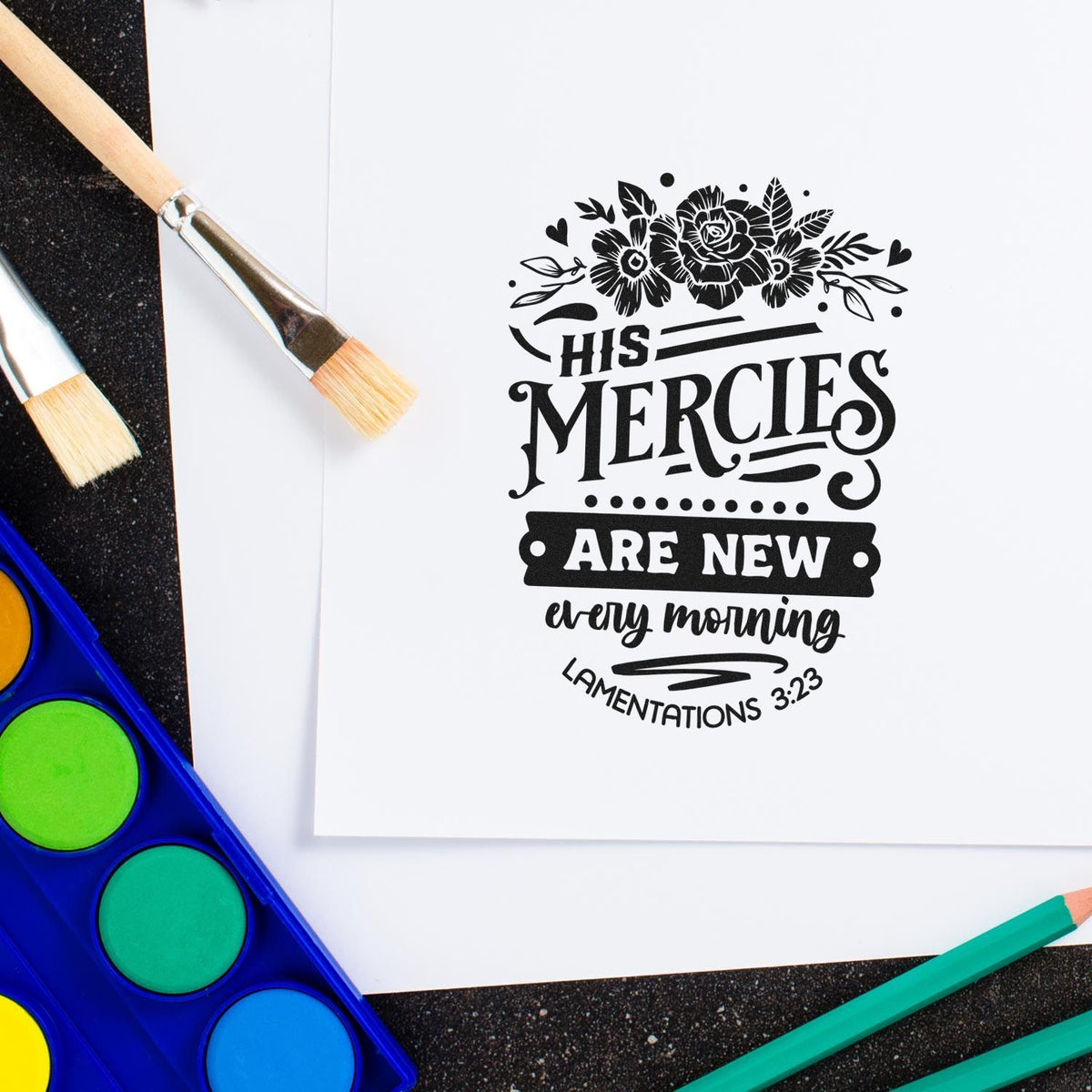 Black floral scripture stamp/seal on white paper, surrounded by paintbrushes and a bright blue watercolor palette.