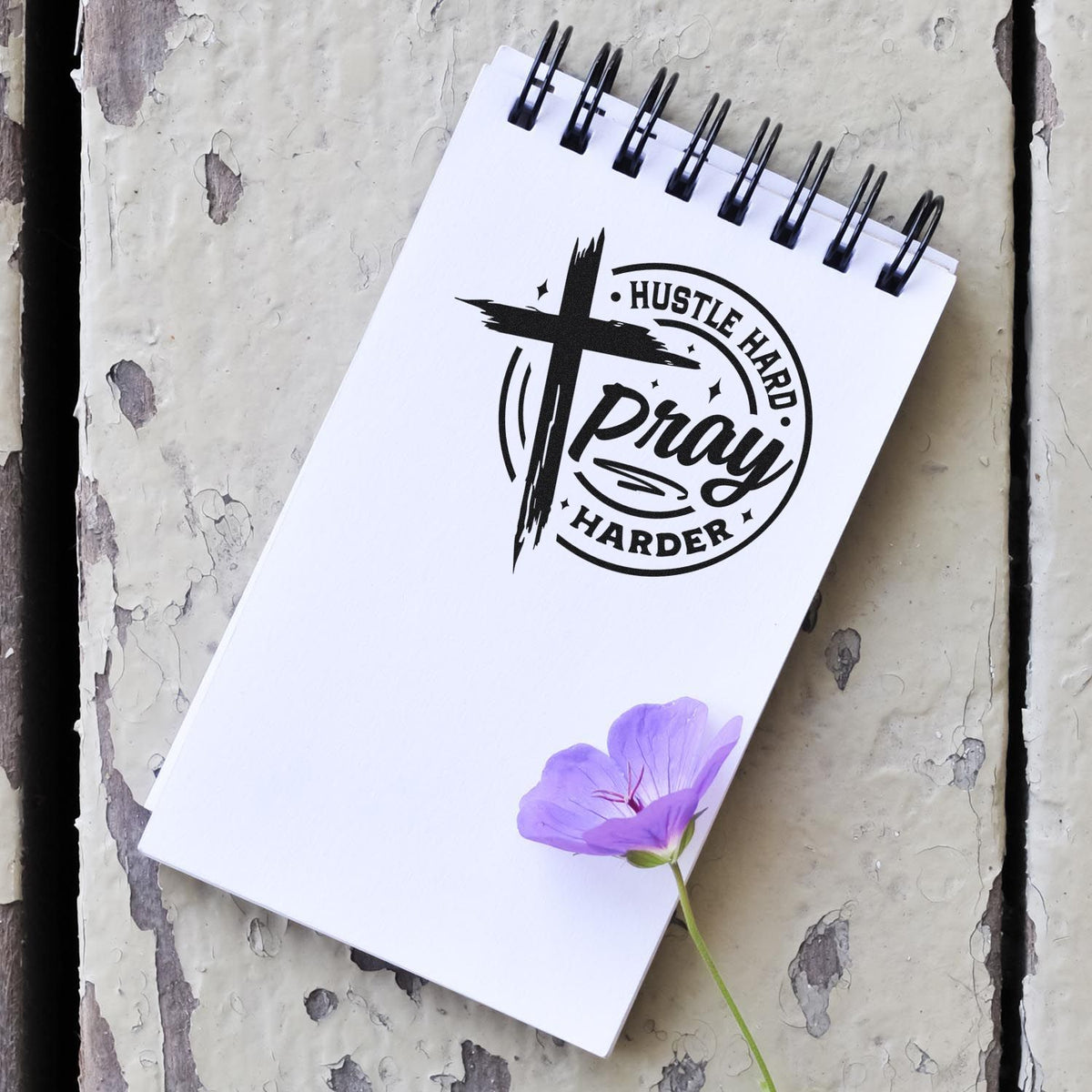 White spiral notepad featuring a black Hustle Hard Pray Harder stamp and seal logo, with a purple flower.