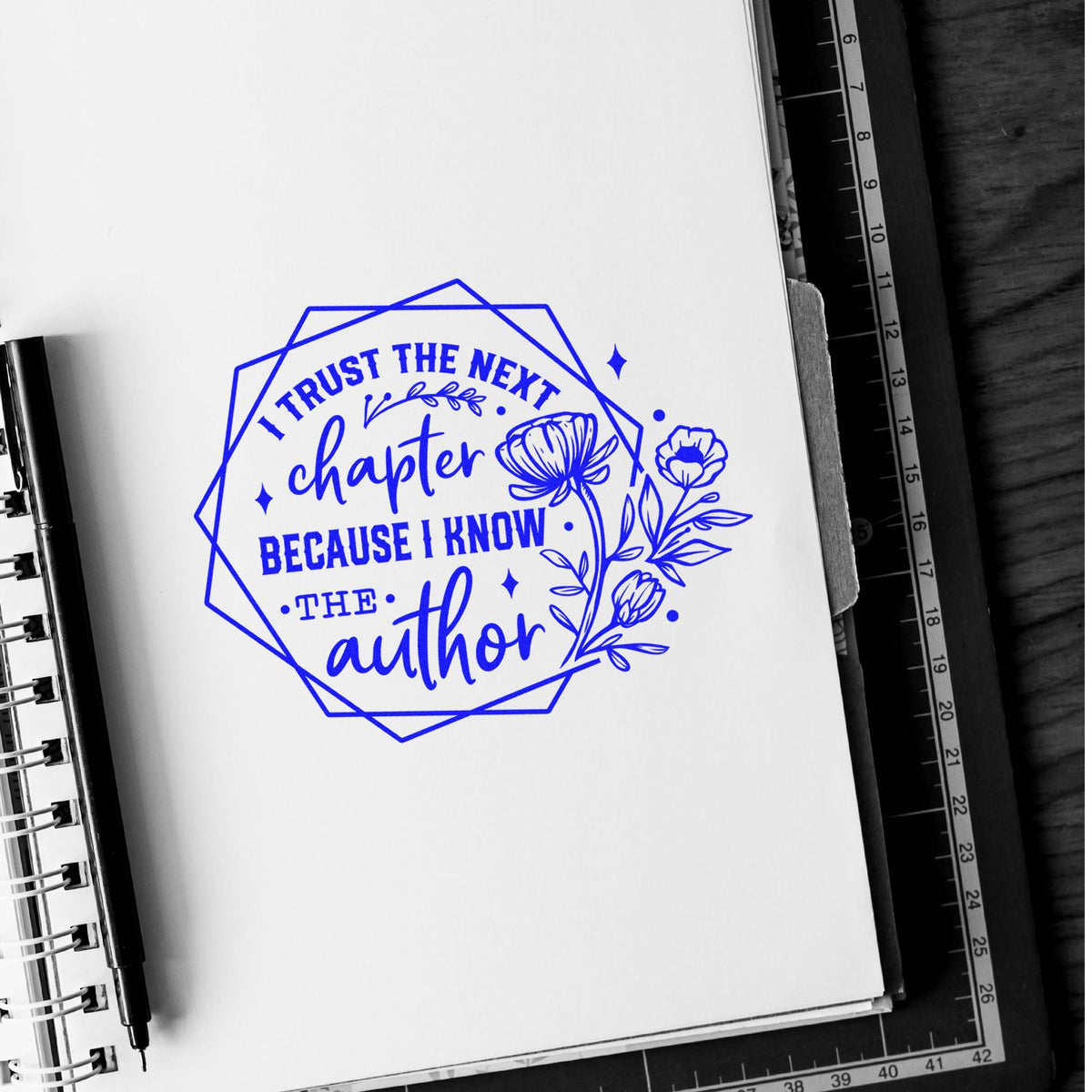 Blue stamp seal reading I trust the next chapter because I know the author, with floral accents on a white notebook.