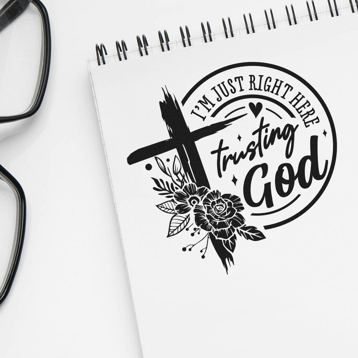 Black faith-themed stamp seal on white spiral notebook, with floral cross, heart and Im just right here trusting God text.