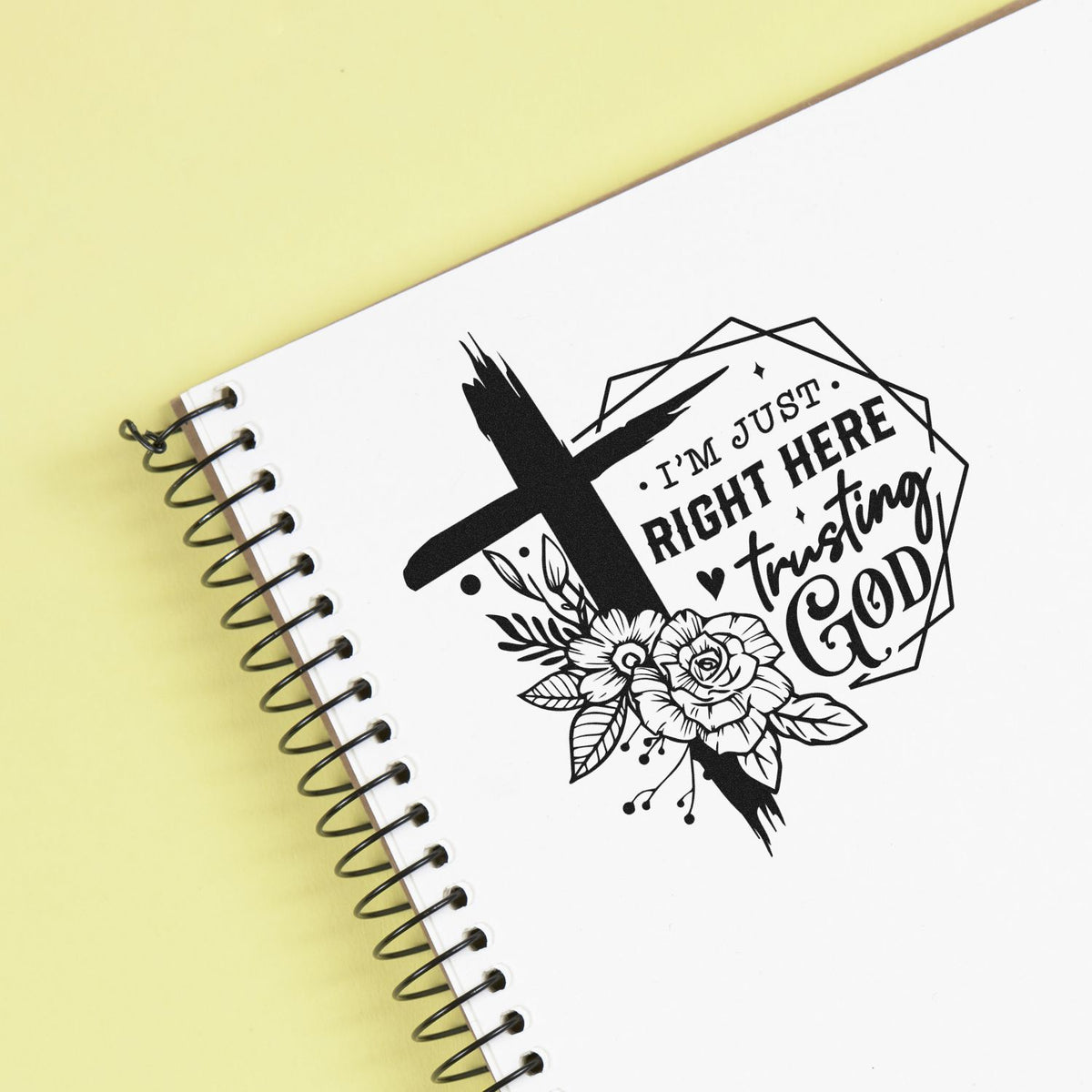 Black faith stamp seal with cross, flowers, and Im just right here trusting God on white spiral notebook, pale yellow background