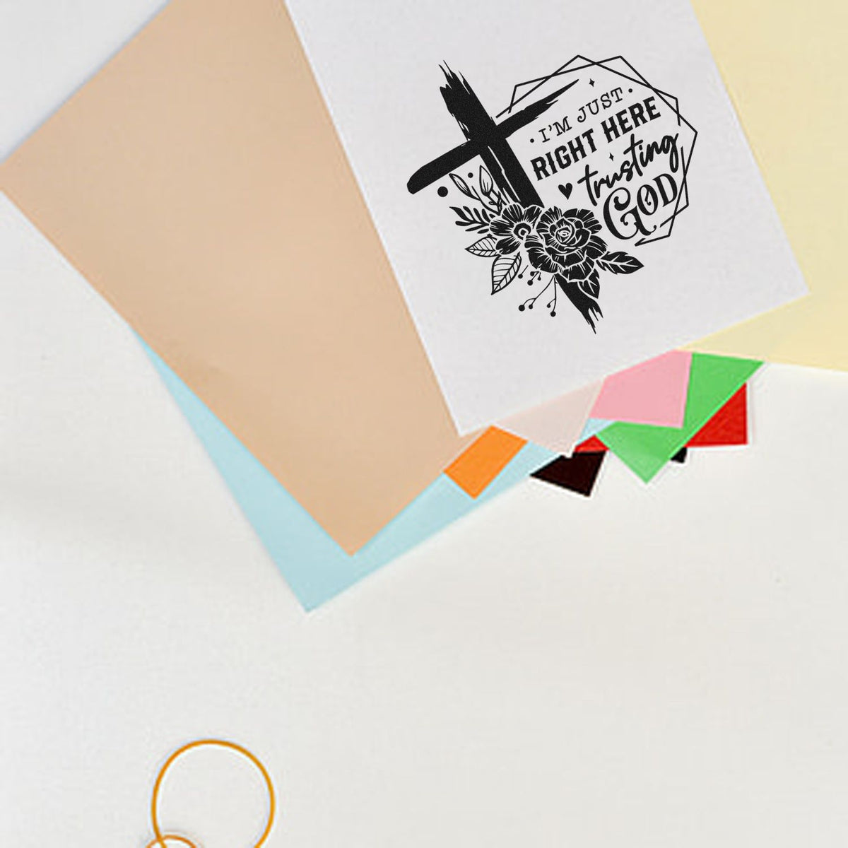 Black religious stamp seal with floral cross on white card, displayed over pastel paper swatches and colorful samples.