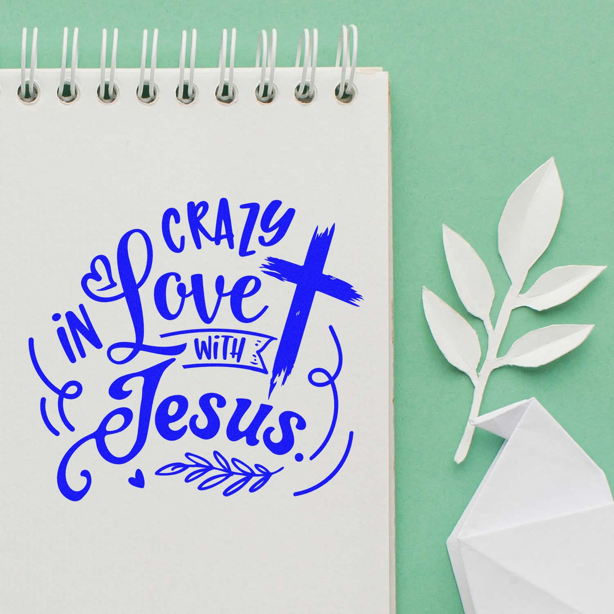 Blue Crazy in Love with Jesus stamp on white spiral notebook, with white paper leaf and origami seal on mint green.