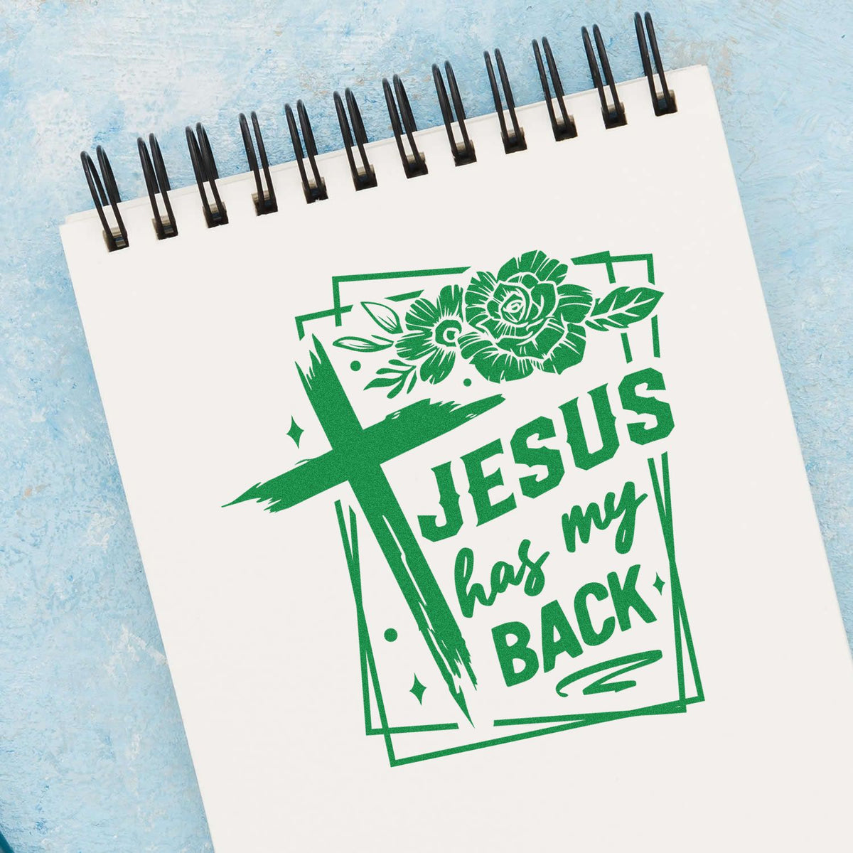 Green Christian stamp design with cross, floral seal, and Jesus has my back on white spiral notebook.