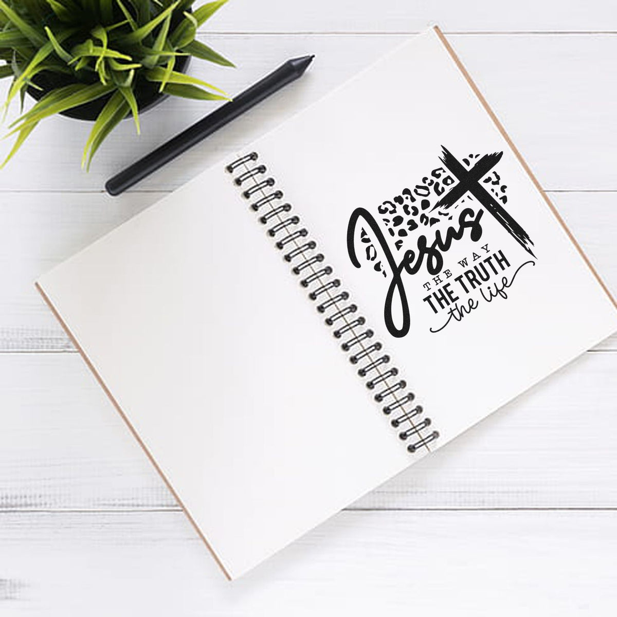 White spiral notebook with black Jesus faith stamp seal design, next to a black pen and green plant.