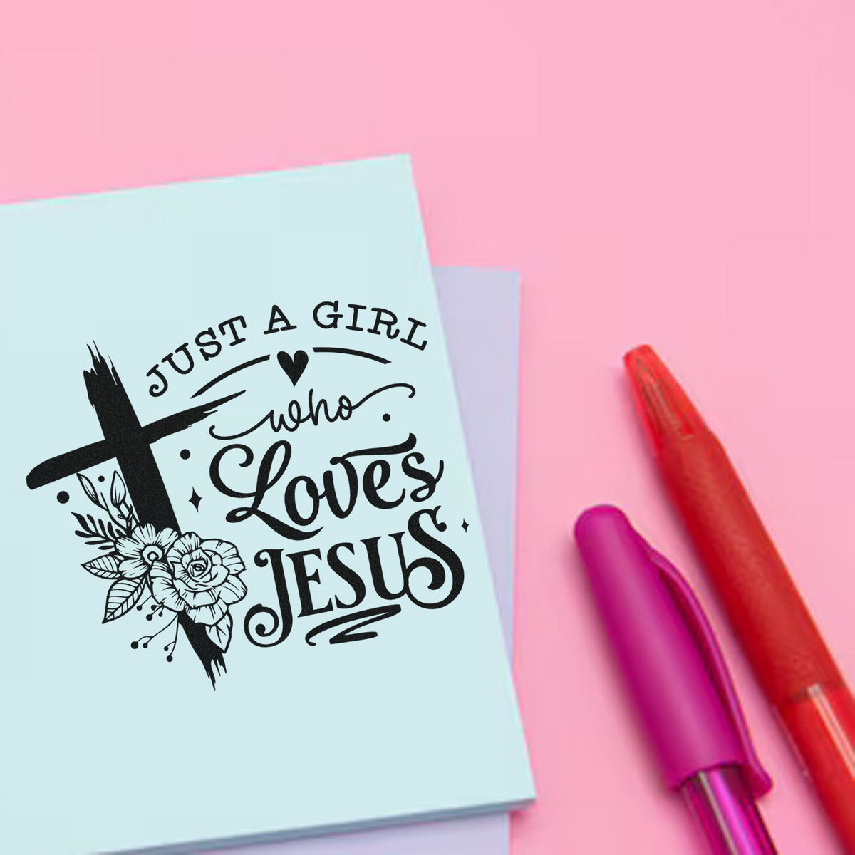 Pastel blue stamp pad with black Christian seal reading Just a girl who loves Jesus, beside red and pink pens on a pink background.