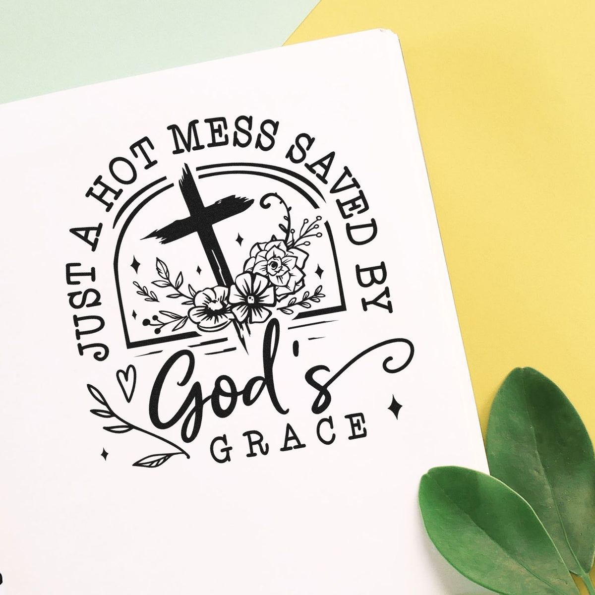 Black stamp seal design with cross and flowers, reading Just a Hot Mess Saved by Gods Grace, on white paper with yellow and green background.