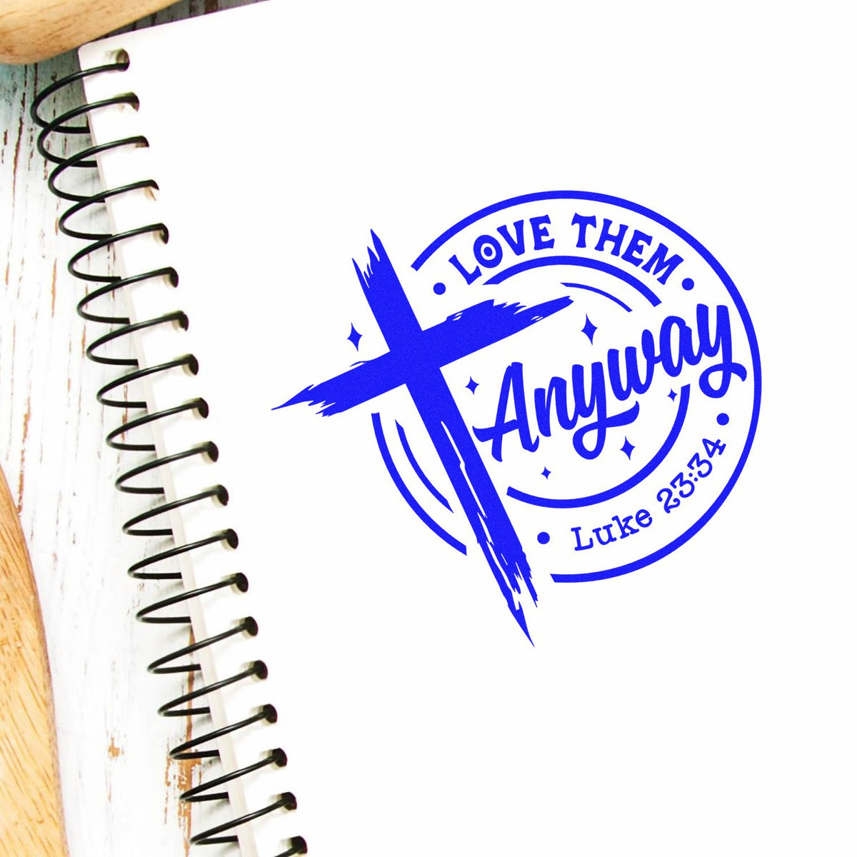 Blue Christian stamp seal with cross and text Love Them Anyway and Luke 23:34 on a white spiral notebook.