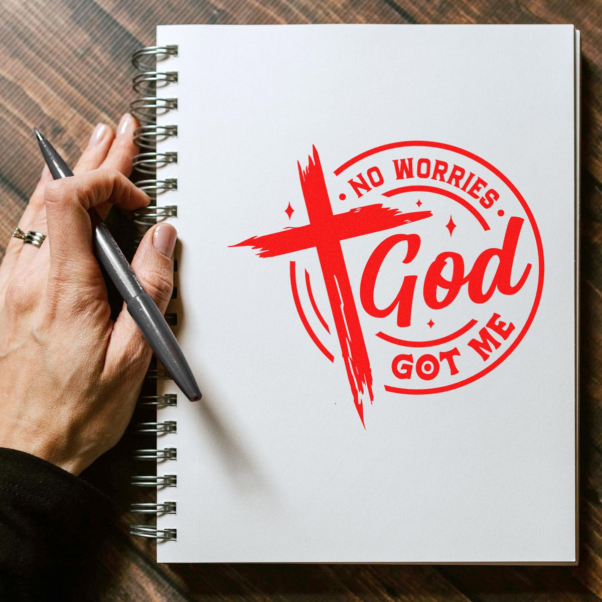Red decorative stamp seal reading No Worries God Got Me on a white spiral notebook, held with a pen.