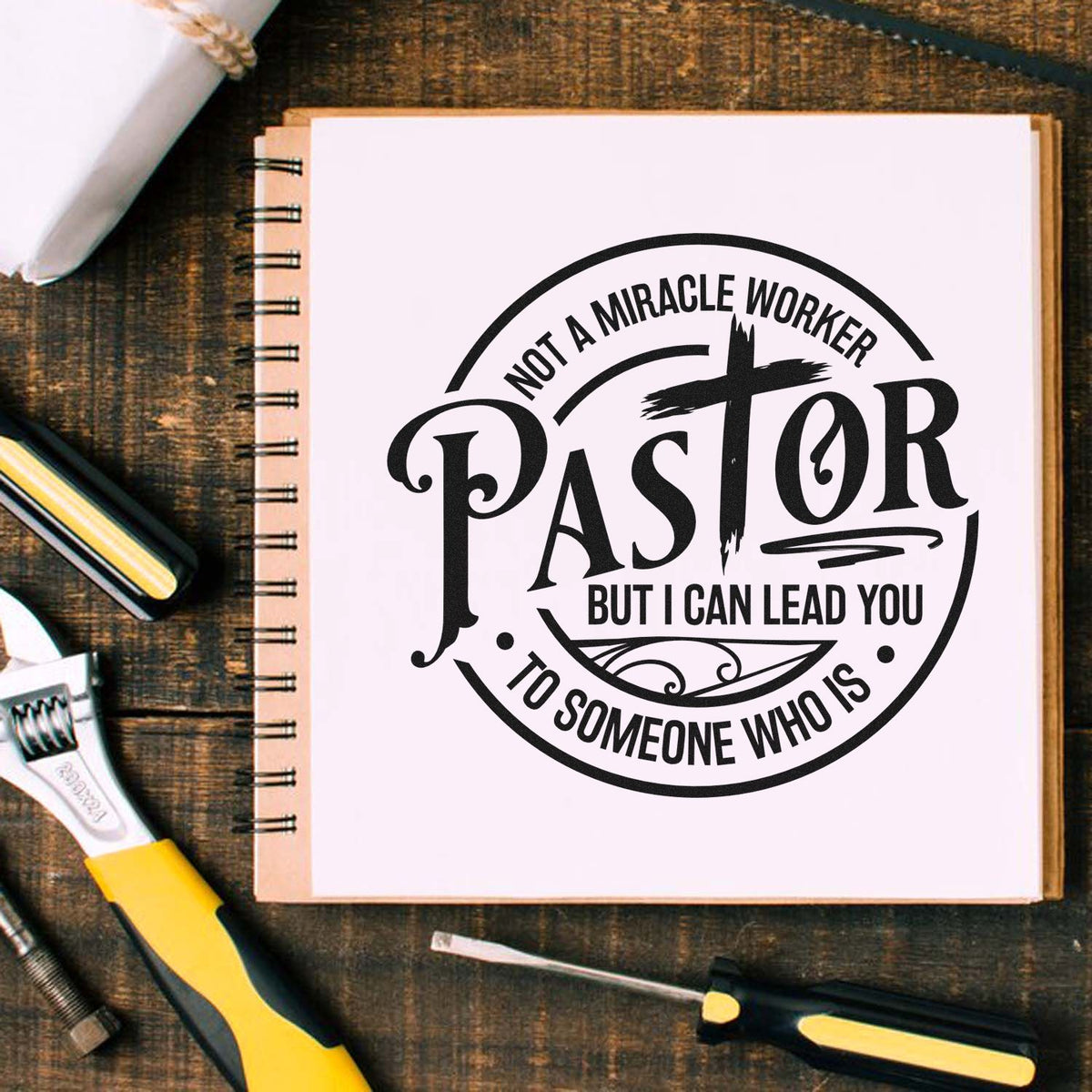 Black pastor stamp seal on a white notebook, with bold circular quote design on rustic wood, plus yellow-handled tools.