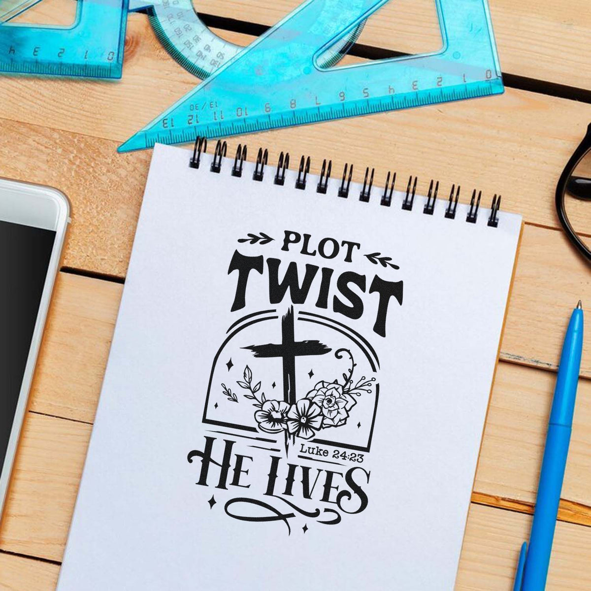 Black Plot Twist He Lives Christian rubber stamp and seal design with cross and flowers on a white notebook.