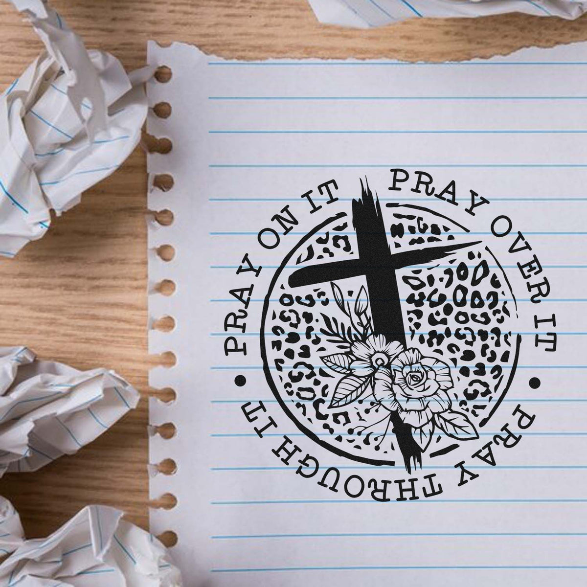 Black prayer cross stamp seal with floral design on white lined notebook paper over a wood desk.