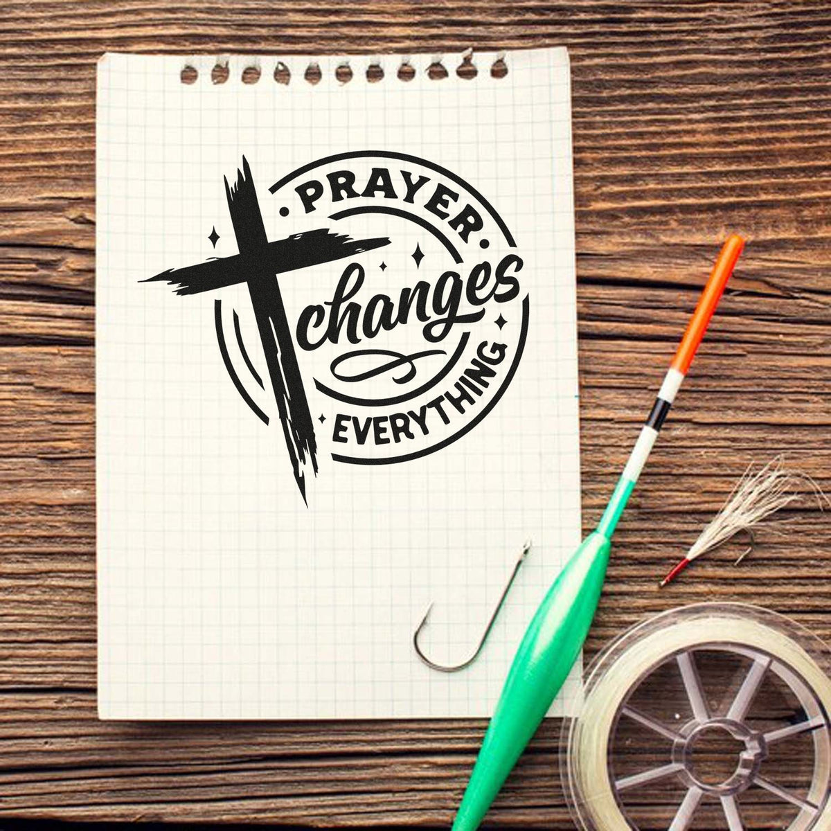 Black prayer changes everything stamp with cross on graph paper notebook, beside fishing hooks and clear seal roll on wood table.