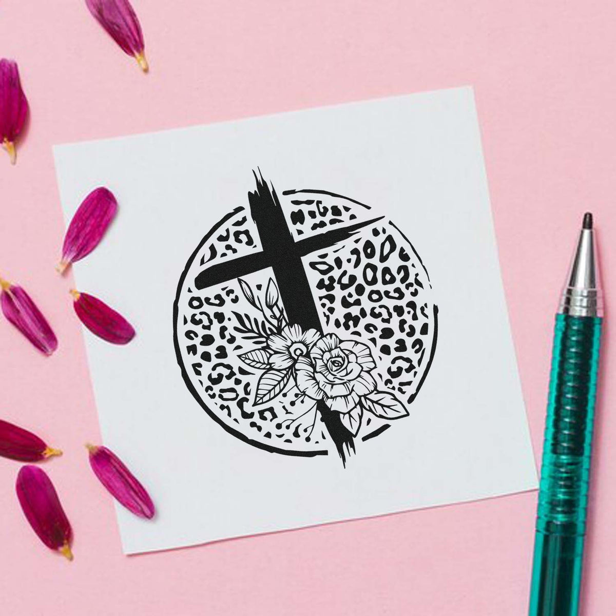 Black cross and floral leopard-print stamp seal on white paper, with pink petals on a pink background and teal pen.