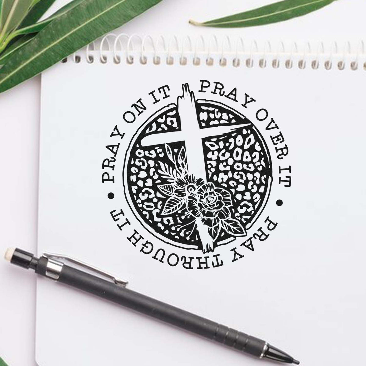 Black Christian prayer stamp and seal design on a white notebook, with a pen and green leaves, featuring a cross and floral details.