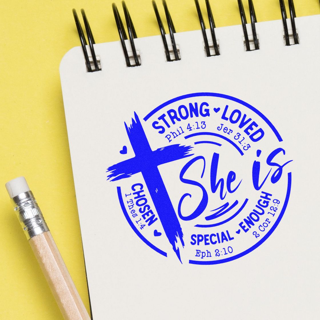 Blue Christian stamp seal reading She is on a white spiral notebook, with yellow background and pencil.