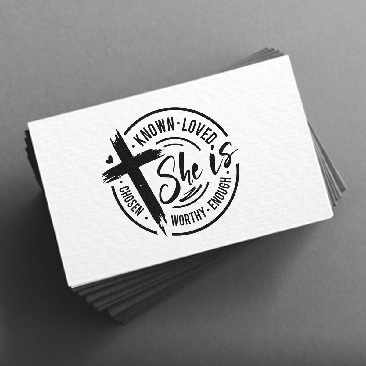 Stacked white stamp cards with black circular She is seal, cross icon, and words known, loved, chosen, worthy, enough on gray background.