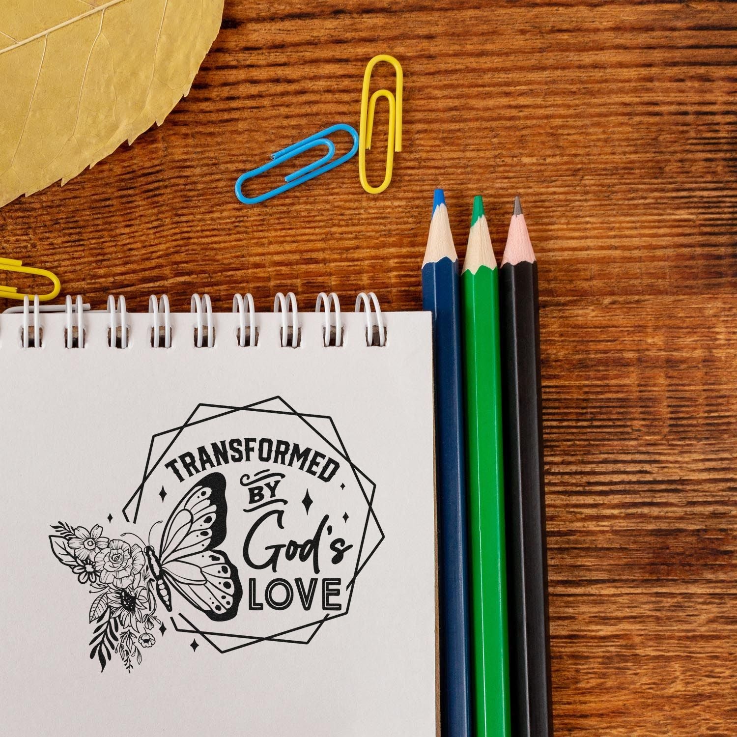 Black custom stamp and seal impression on white notebook, with butterfly, floral art, and blue, green, black pencils.
