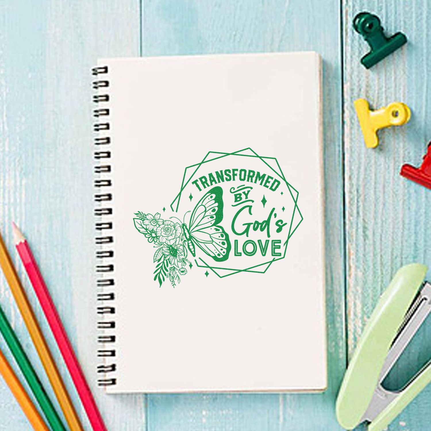 White spiral notebook with green butterfly stamp seal reading Transformed by Gods Love, on blue wood with colorful clips.