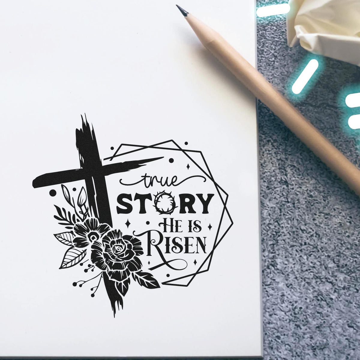 Black stamp and seal design with cross, flowers, and True Story He Is Risen on white paper beside a pencil.