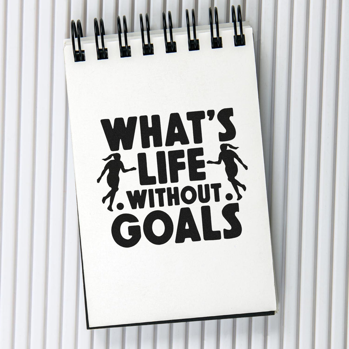 Black stamp-style seal reading WHATS LIFE WITHOUT GOALS on a white spiral notebook, with running silhouettes and bold black text.