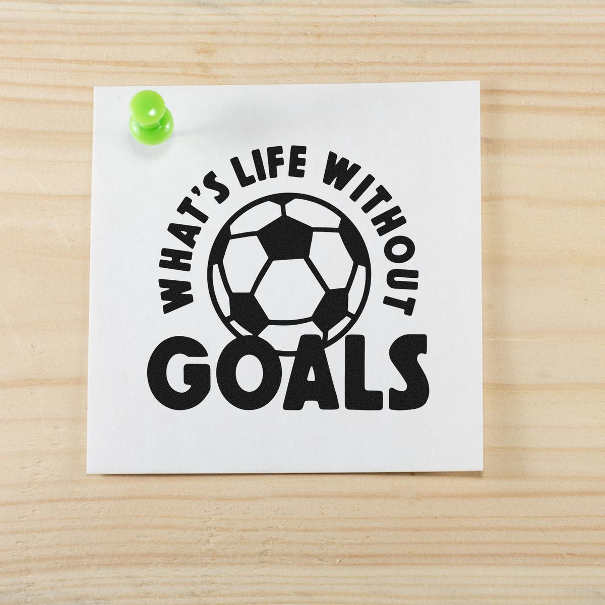 Black soccer goals stamp seal on white card, pinned with a green pushpin, on a light wood background.