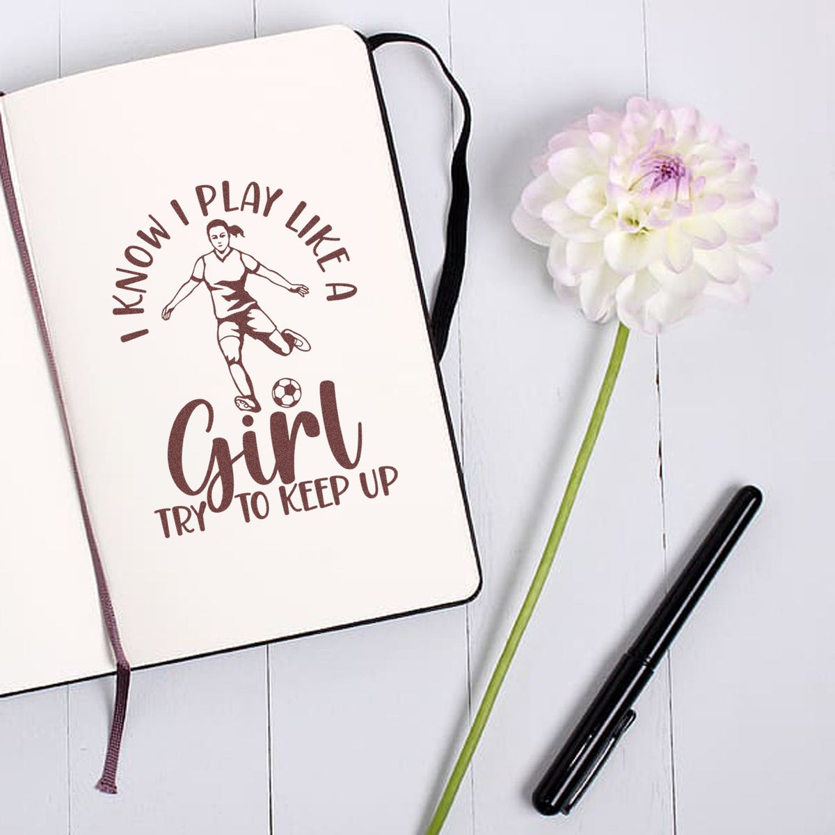 Brown soccer girl stamp design on cream notebook, with black pen and white flower, reading I know I play like a girl.