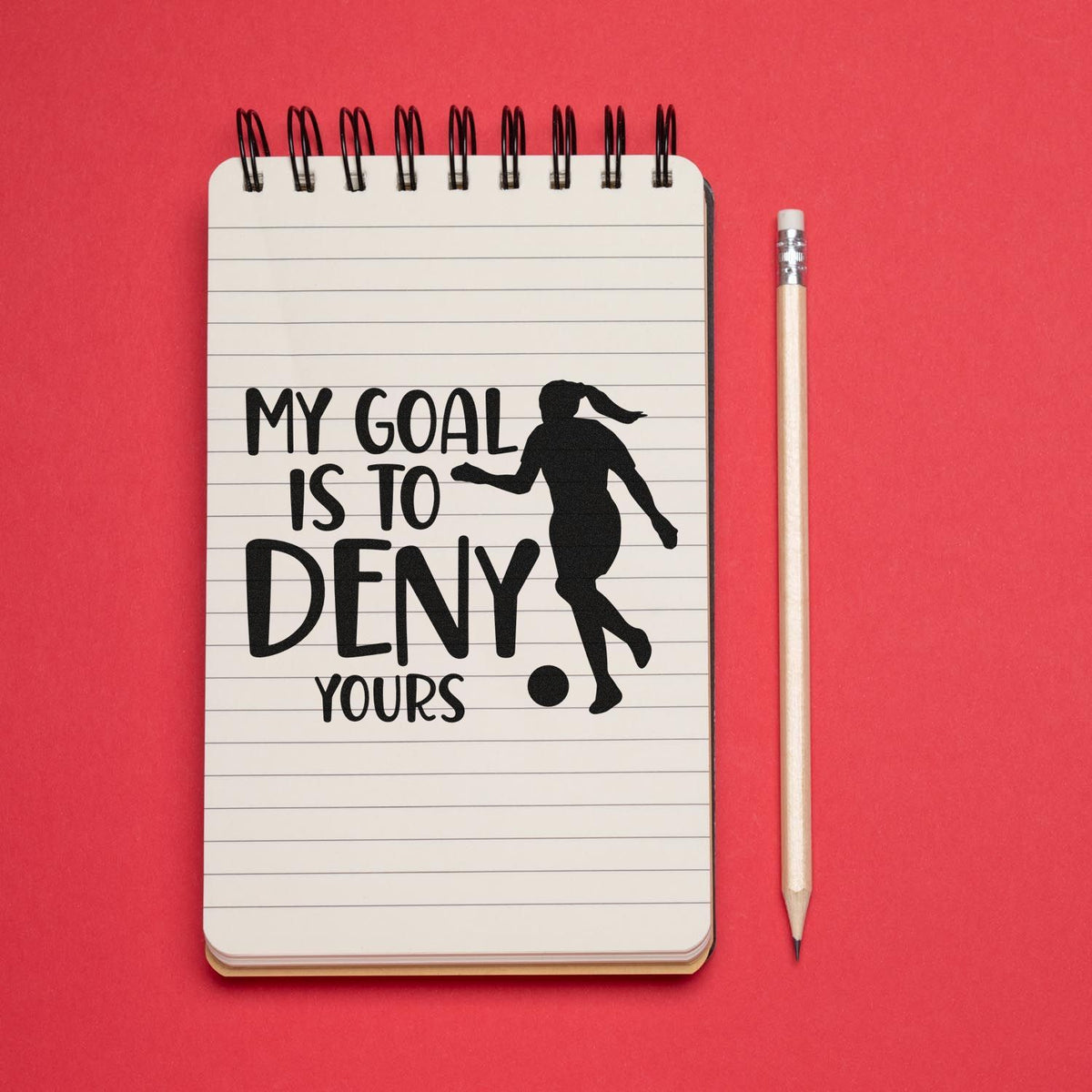 Black rubber stamp seal reading My goal is to deny yours on a white spiral notebook, with soccer silhouette, red background and pencil.