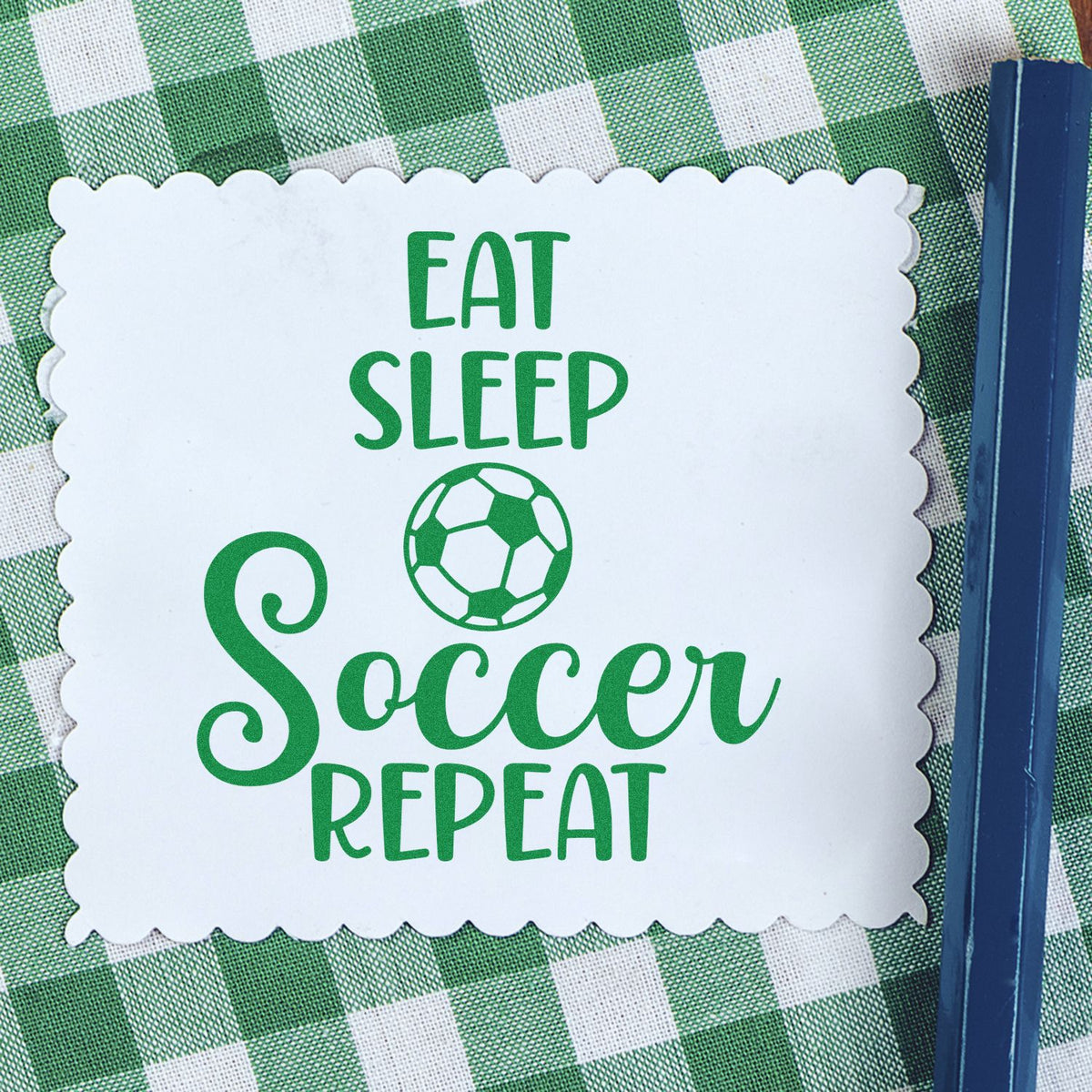 Green soccer stamp seal reading Eat Sleep Soccer Repeat on white scalloped paper over green gingham.