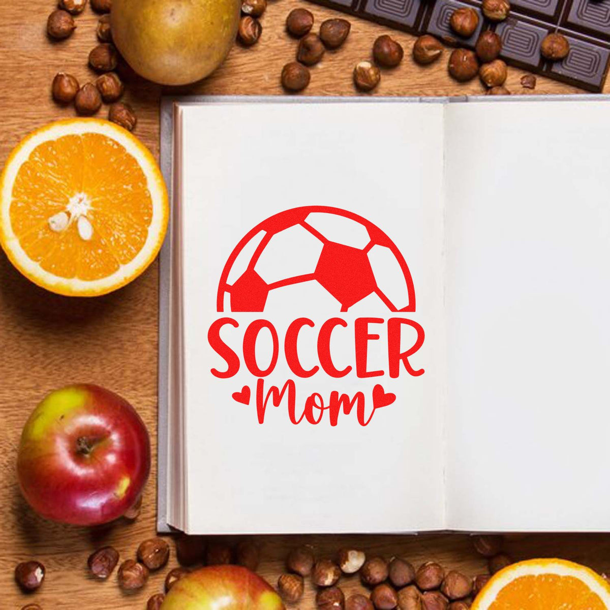 Red soccer mom stamp seal with soccer ball and hearts on white paper, surrounded by oranges, apple, and coffee beans.