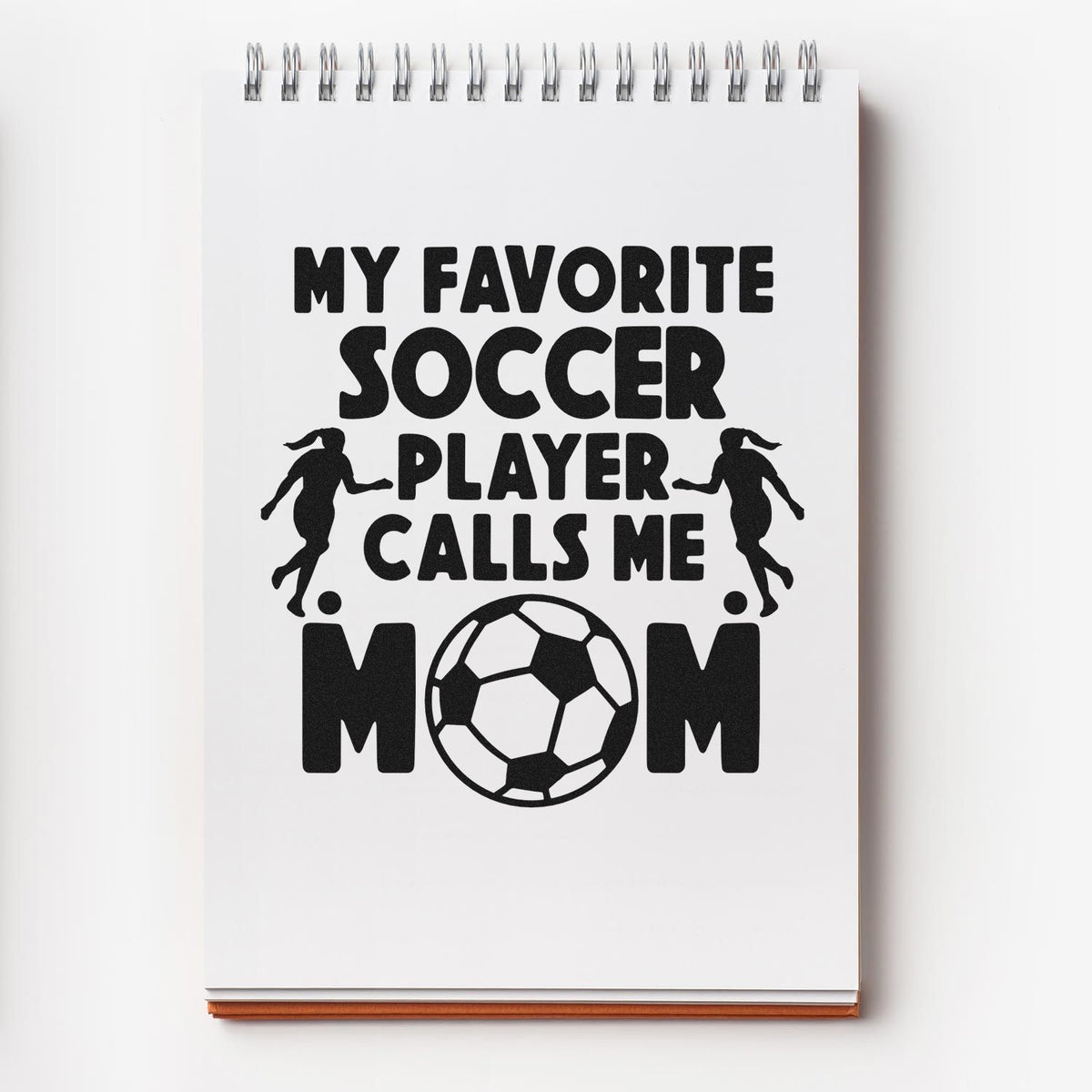 Black stamp and seal design on white spiral notebook, reading My favorite soccer player calls me mom with soccer ball and player silhouettes.