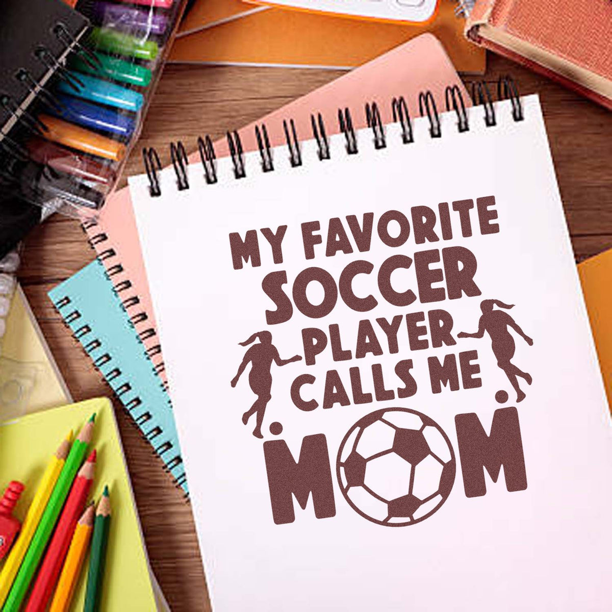 Brown custom stamp/seal on white spiral notebook reads My favorite soccer player calls me mom, with soccer ball and players.