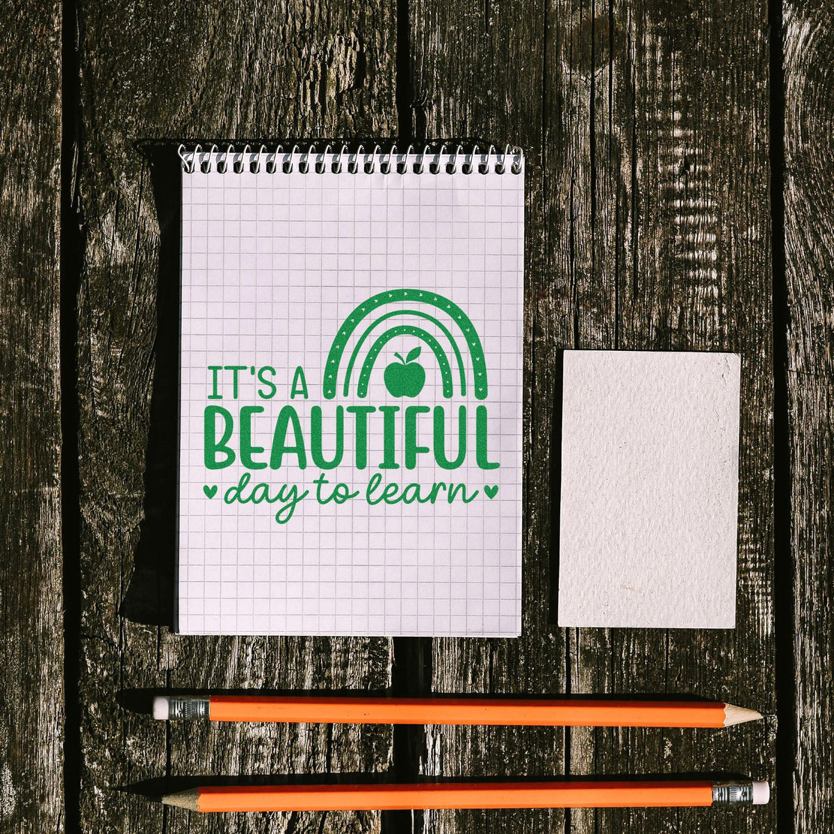 Green stamp and seal set on grid notepad reading Its a beautiful day to learn, with blank card and orange pencils on dark wood.