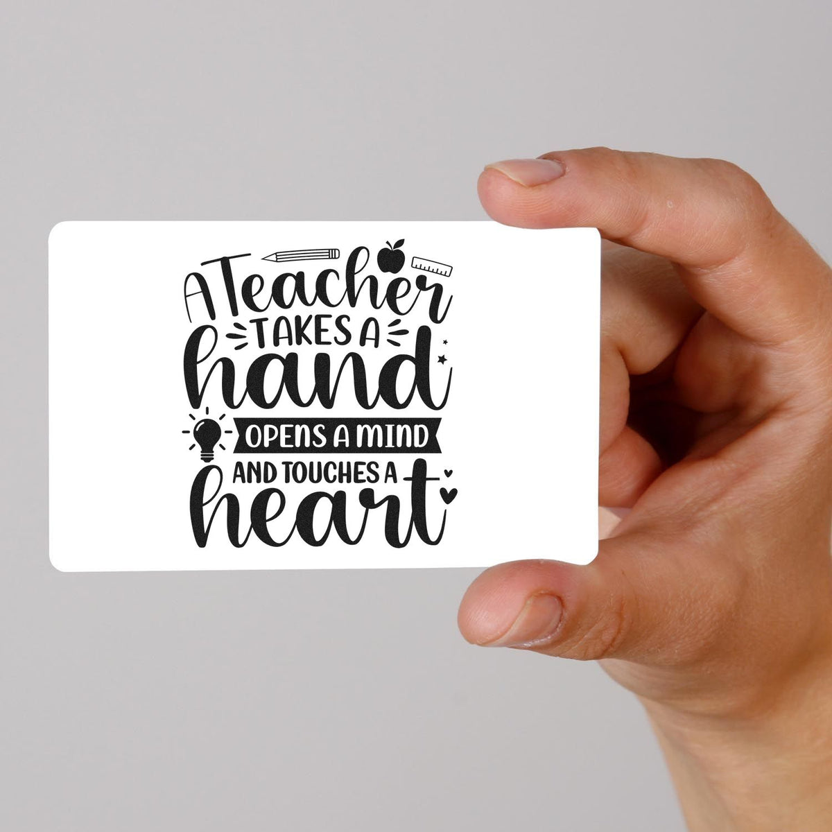 Hand holding white stamp seal card with black teacher quote, pencil, apple and ruler icons on a gray background.