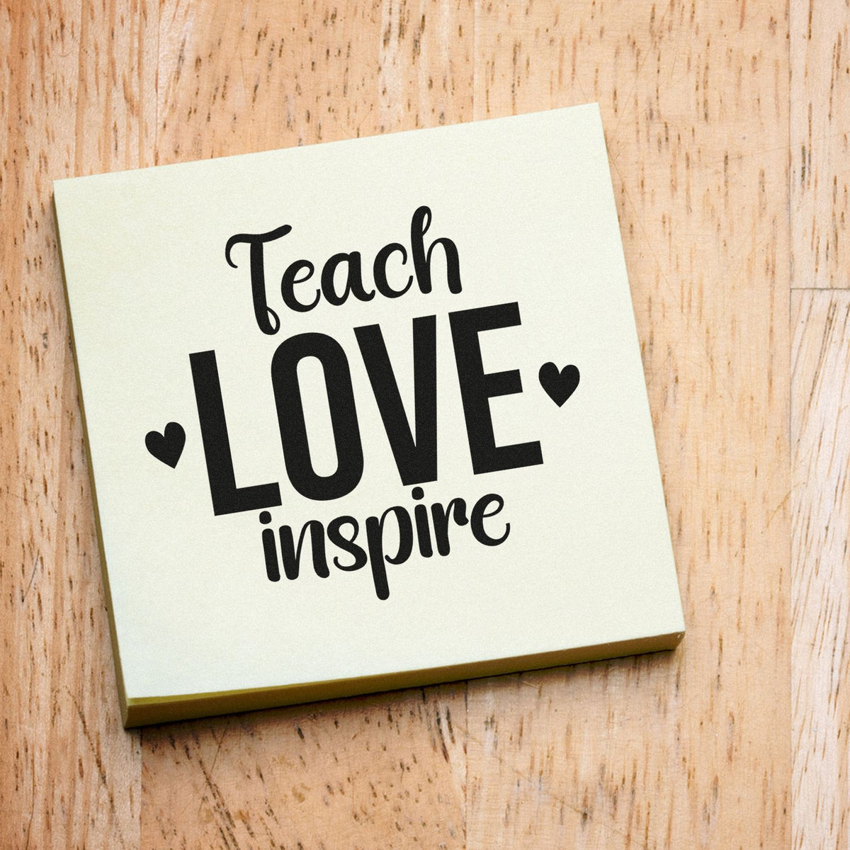 Square cream teacher stamp with black Teach LOVE inspire text, heart icons, on a light wood background.