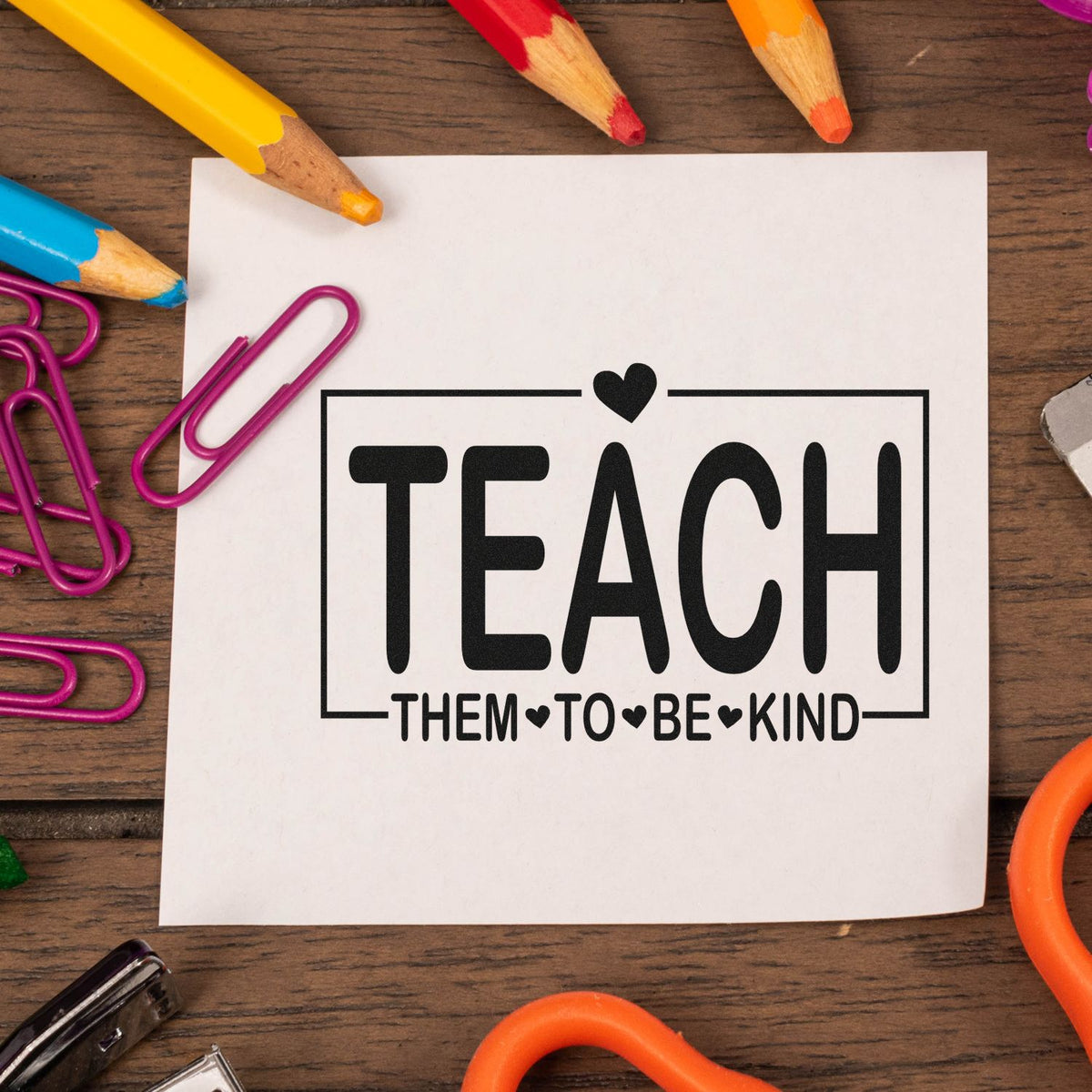 Black teaching stamp seal reading "Teach them to be kind" on white paper, with colorful pencils and magenta paper clips.