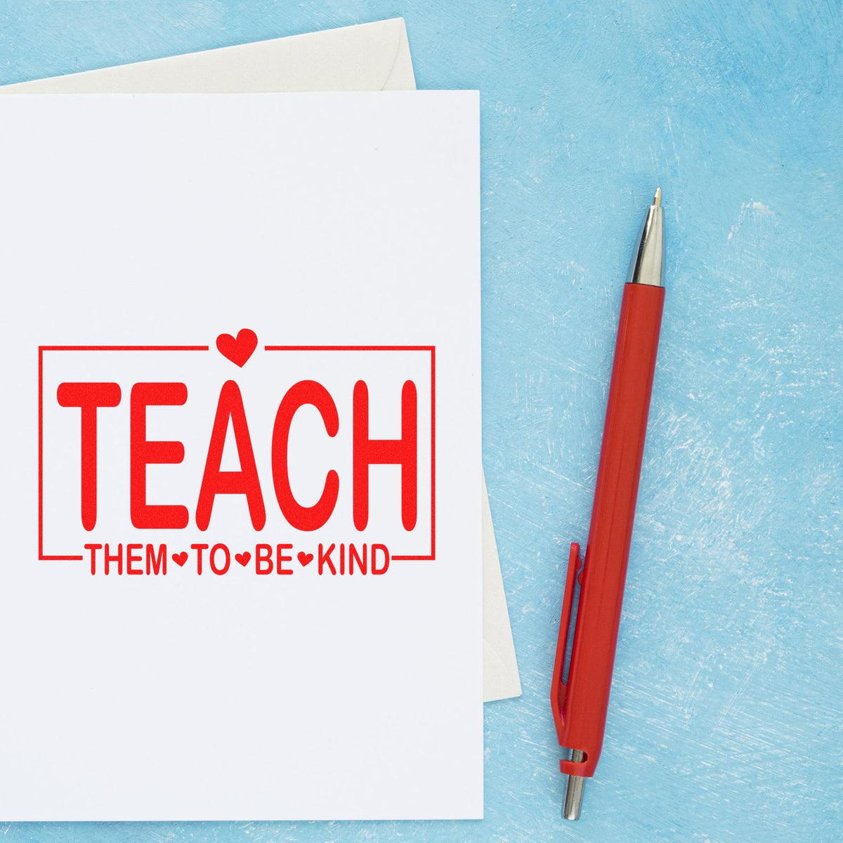 Red teacher stamp and seal reading Teach Them to Be Kind beside a red pen on a blue background.