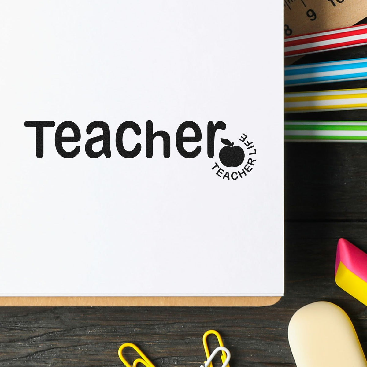 Black teacher stamp and seal with apple icon on white paper, beside colorful stationery on a dark desk.