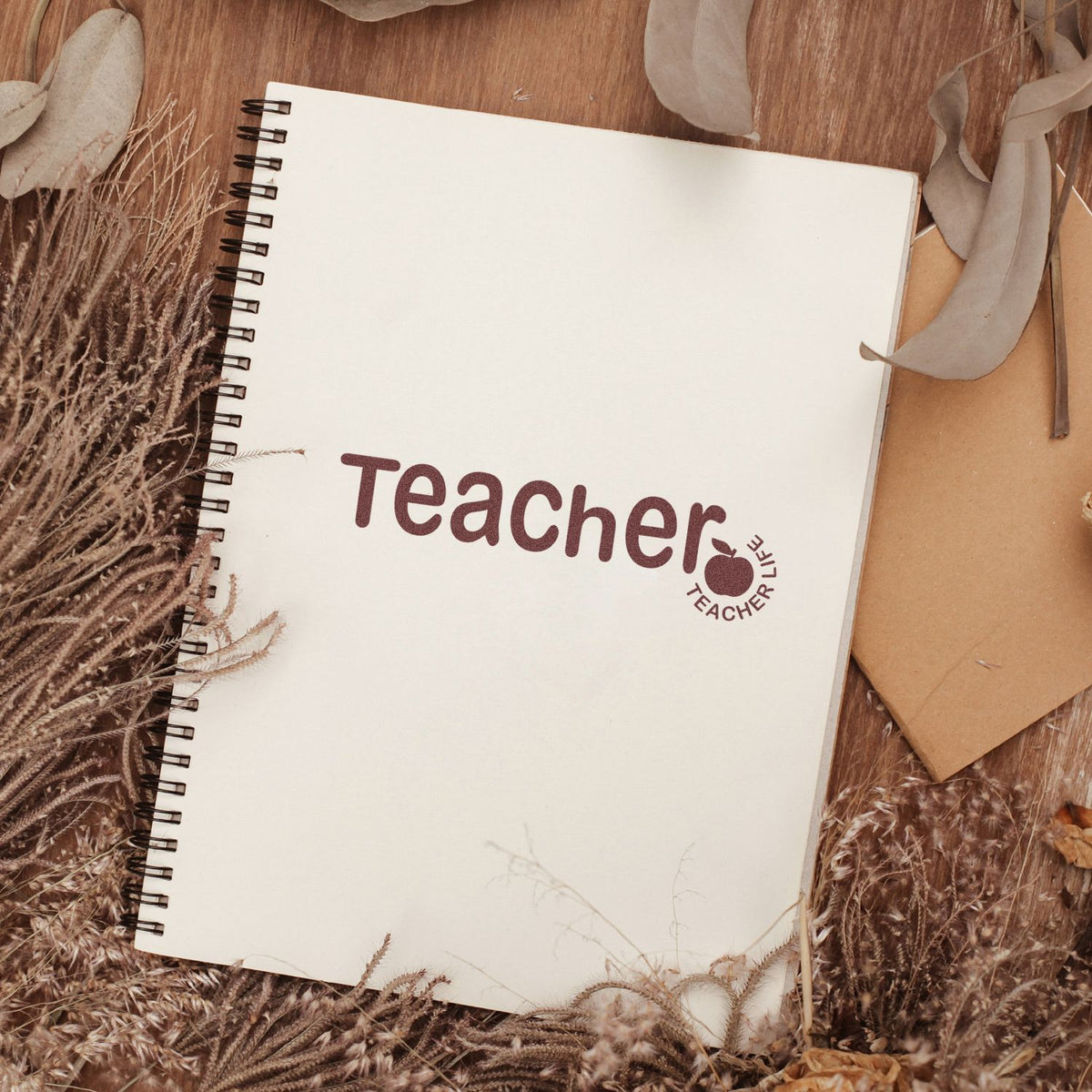 Cream spiral notebook with maroon Teacher stamp and matching Teacher Life seal on a warm brown rustic background.
