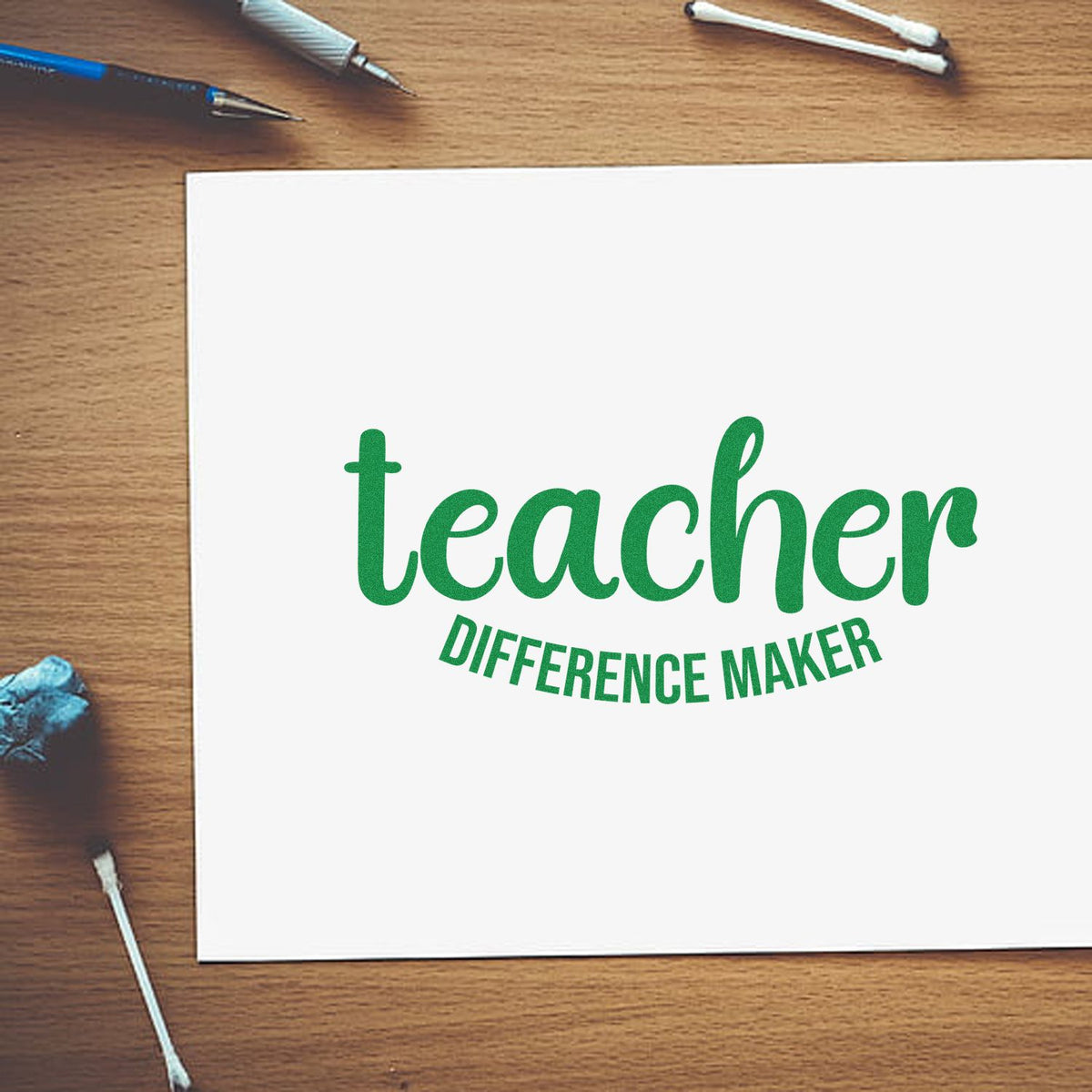 Green teacher difference maker stamp seal on white paper, with desk tools on a wood tabletop.