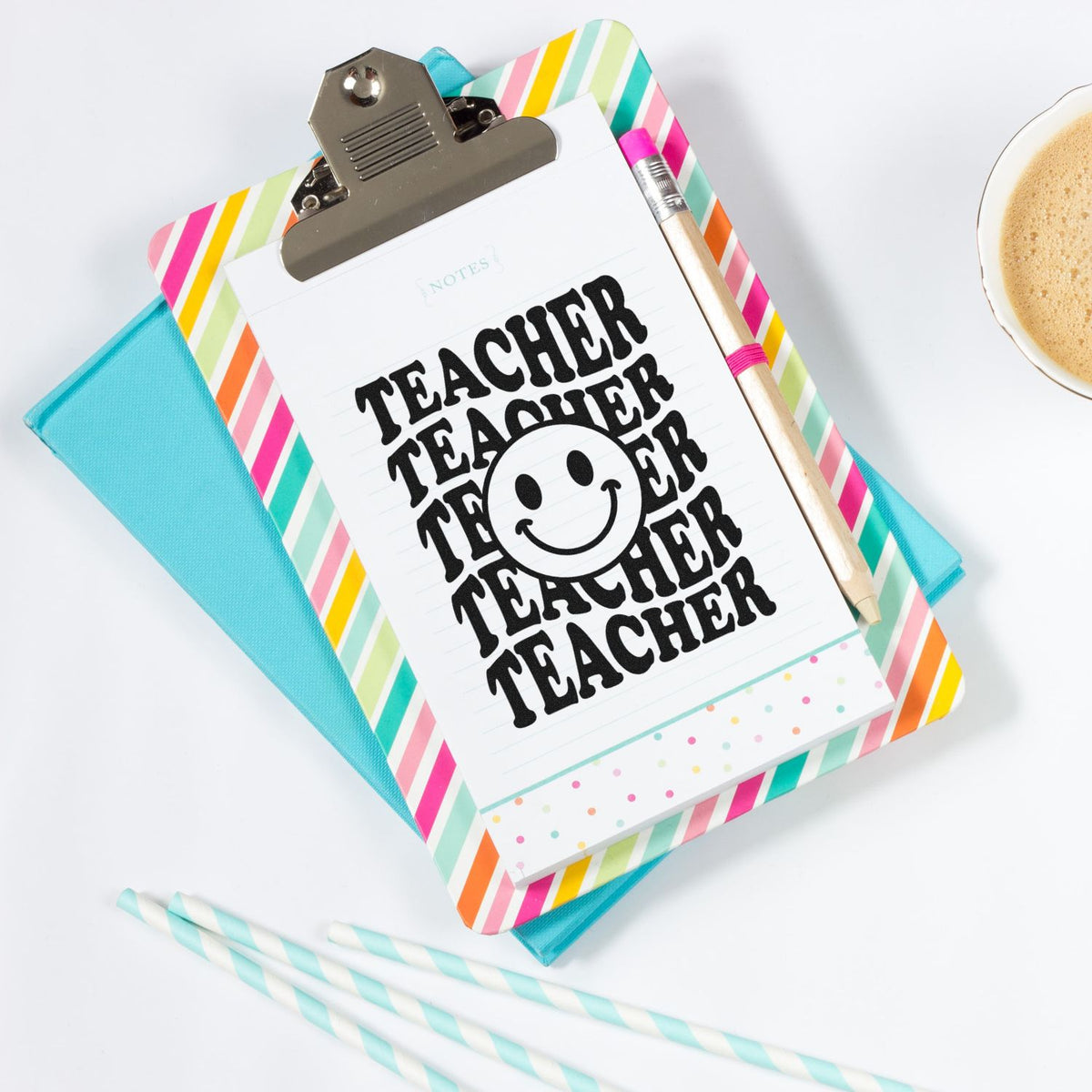 Black teacher stamp with smiley face on rainbow-striped clipboard, with turquoise folder, pencil, and coffee cup.