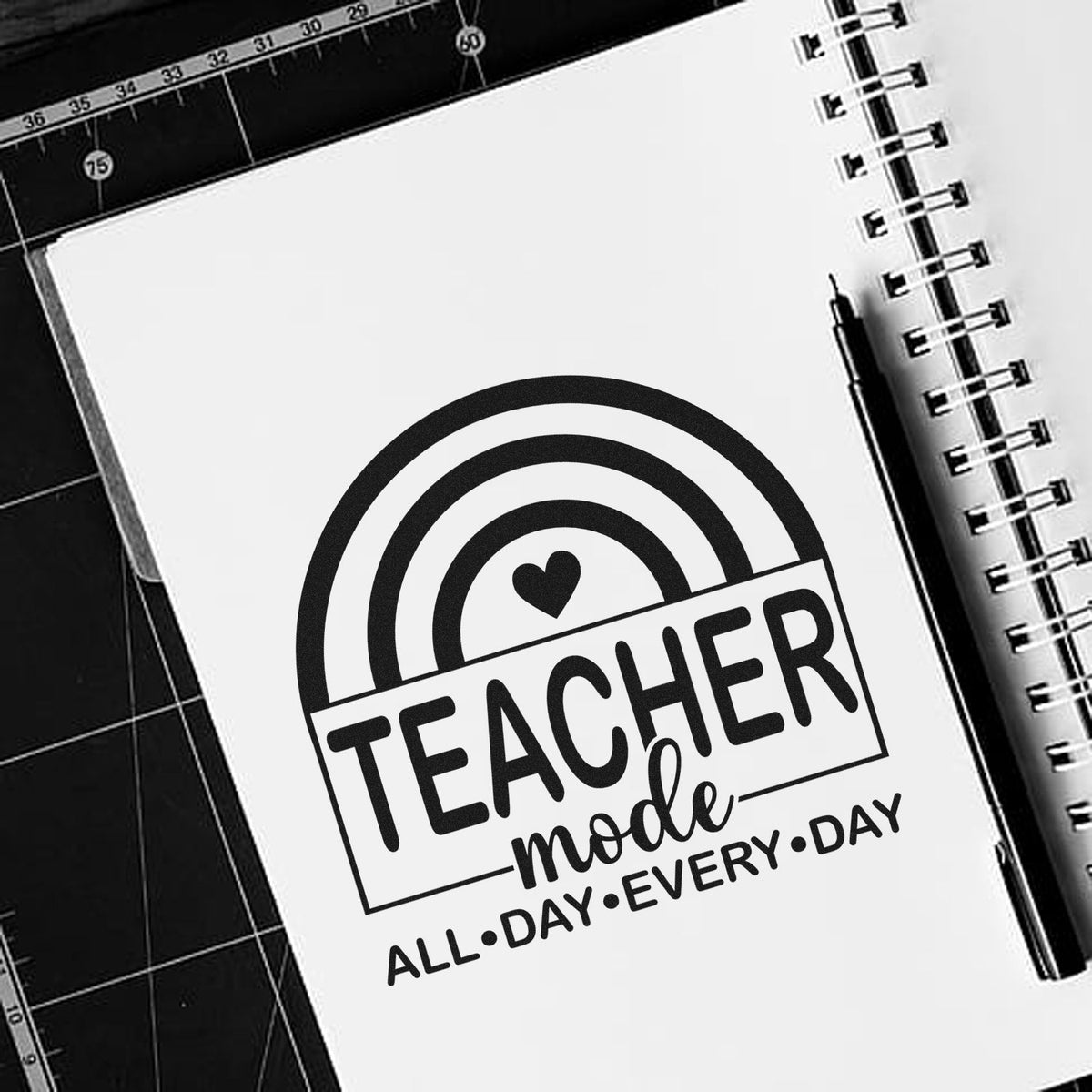 Black-and-white teacher-mode stamp and seal design with rainbow, heart, and notebook spiral on a white page.