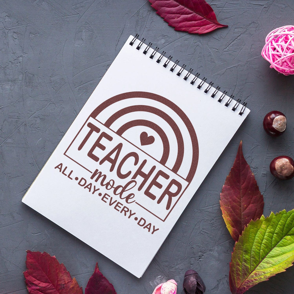 Brown teacher stamp seal on white spiral notebook with rainbow heart, on gray background with burgundy and green fall leaves.