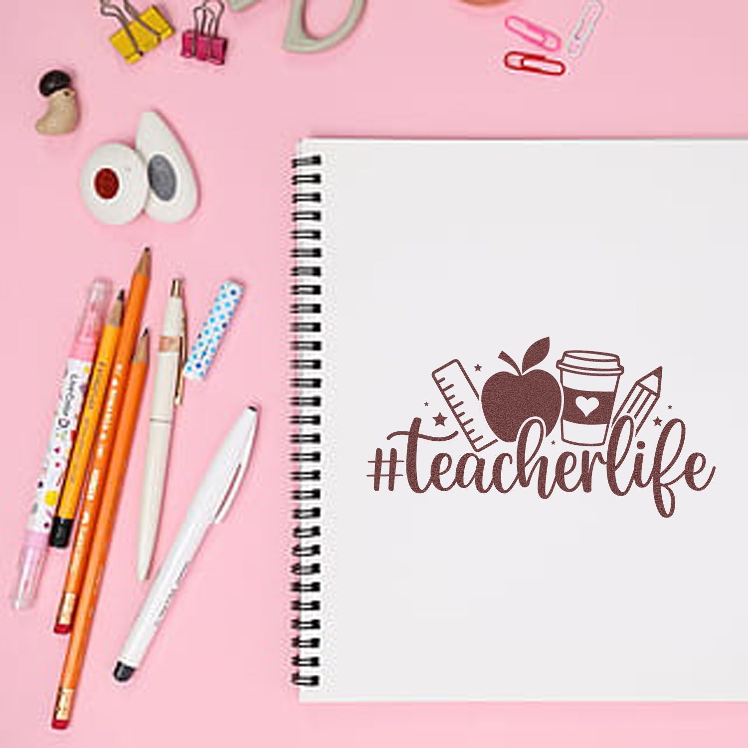 White spiral notebook with a brown teacherlife stamp, apple, ruler, coffee cup and pencil on pink, surrounded by pens and clips.