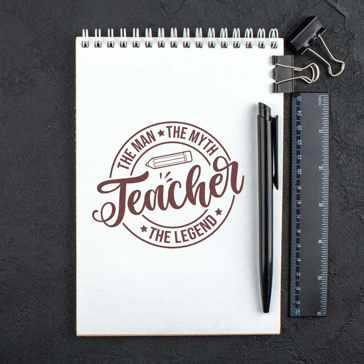 Brown teacher stamp seal on white spiral notebook, with pencil icon, ruler, pen, and binder clips on a dark background.