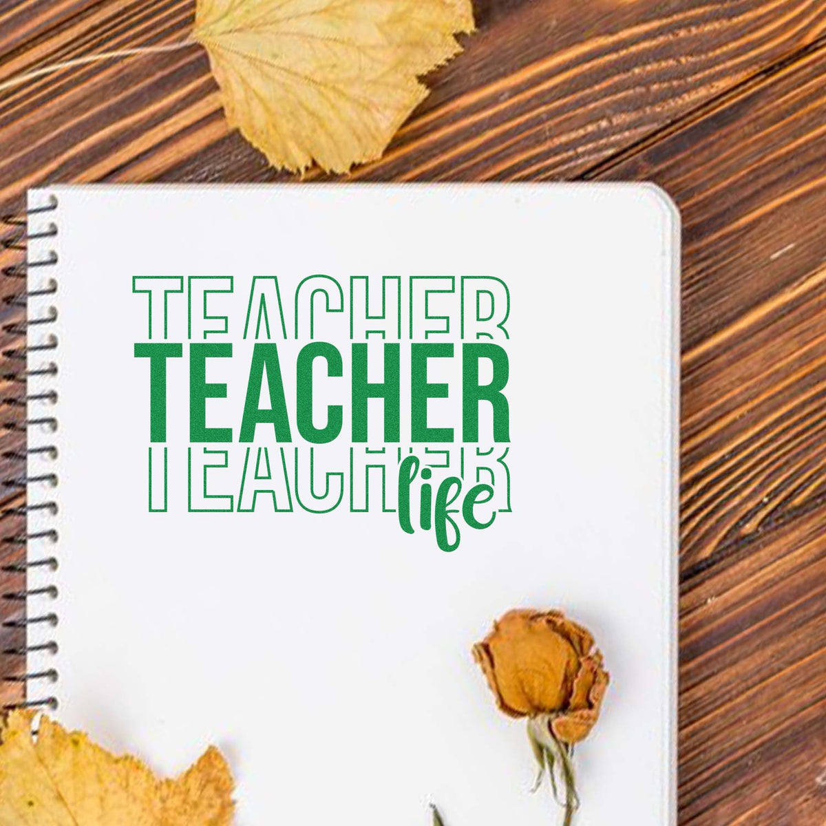Green Teacher Life stamp/seal design on white spiral notebook, with autumn leaves on a wood table.