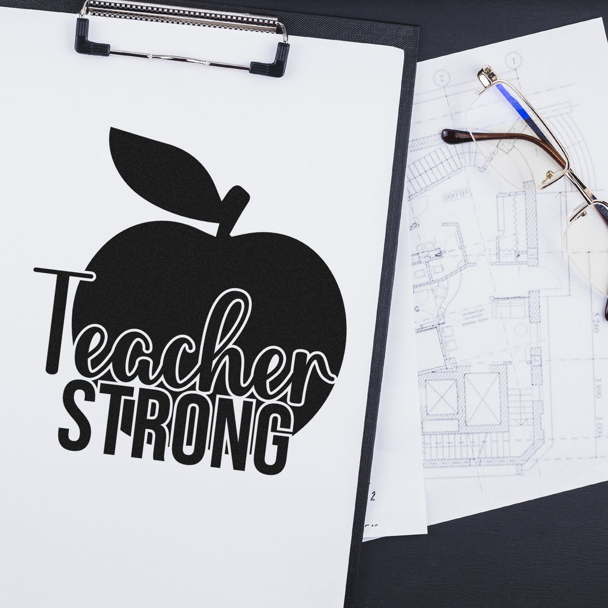Black custom teacher stamp and seal with apple logo on white clipboard beside eyeglasses and blueprint sketches.