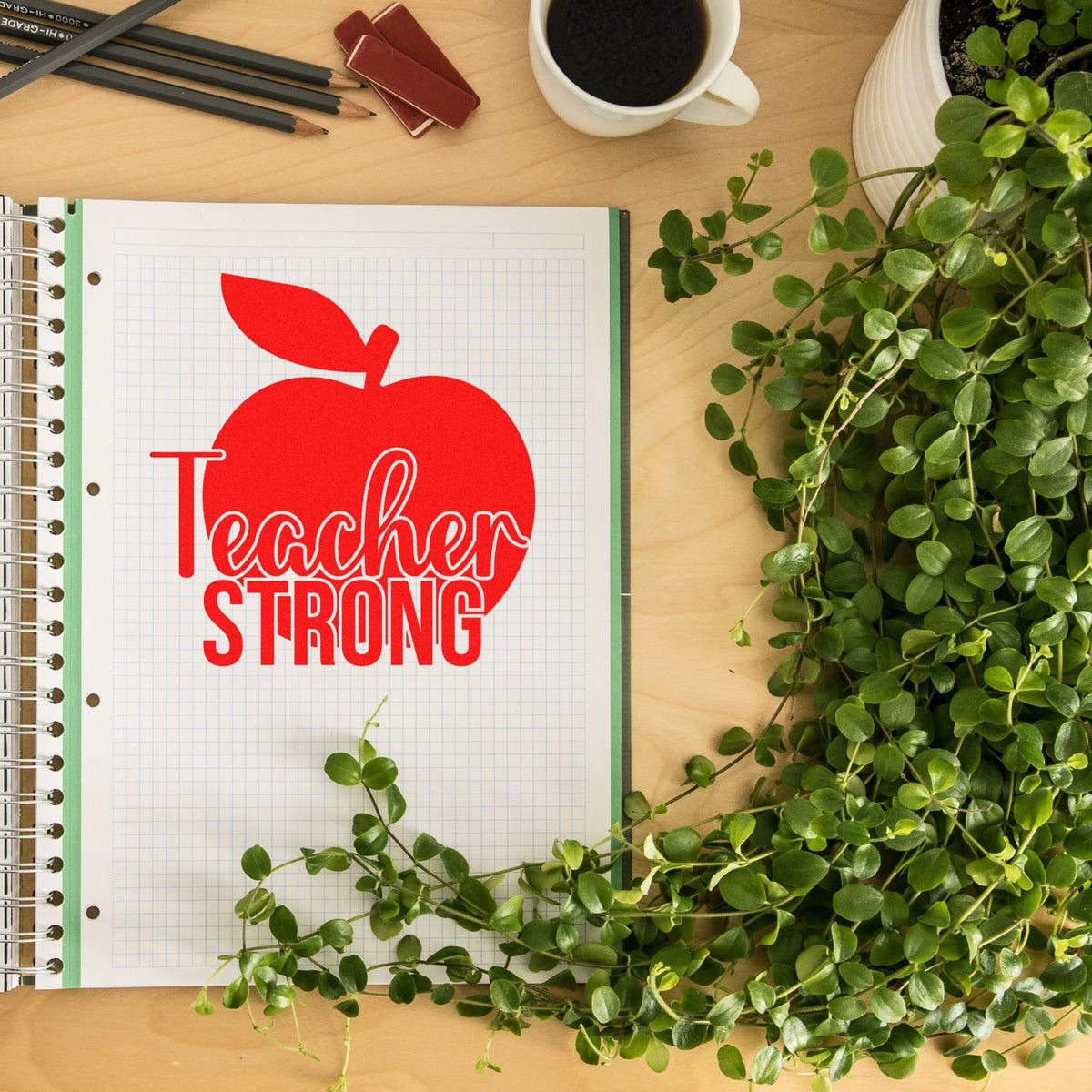 Red Teacher Strong apple stamp/seal on grid paper, surrounded by green plant and desk items.